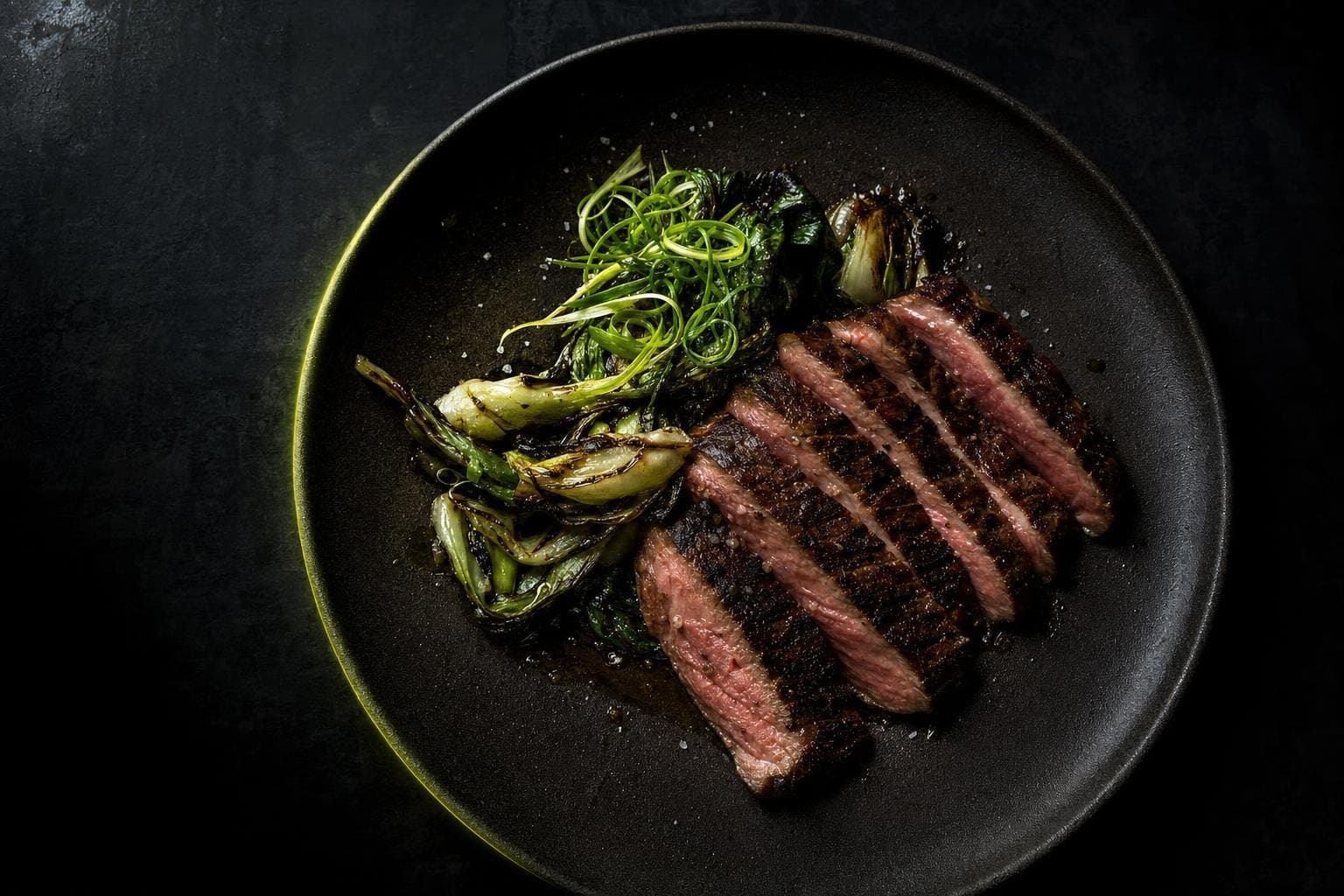 A protein-forward restaurant plate of sliced seared steak with greens on dark ceramic